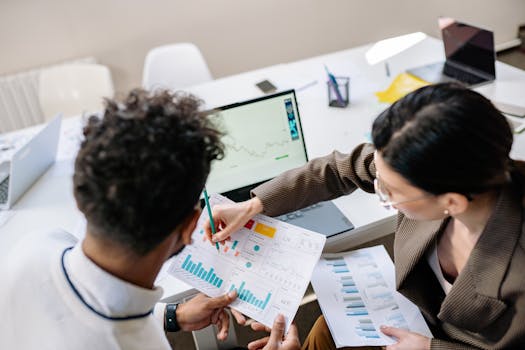 pexels-photo-7693745-7693745 Two colleagues in a meeting room discussing financial charts and graphs on a laptop and paper.